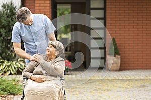 Male helping happy elderly woman in the wheelchair in front of house