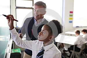 Male High School Teacher With Female Student Wearing Uniform Using Interactive Whiteboard During Lesson