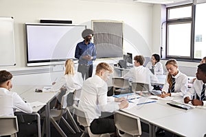 Male High School Teacher Standing Next To Interactive Whiteboard And Teaching Lesson To Pupils Wearing Uniform