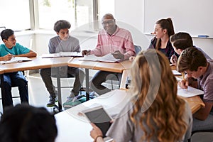 Male High School Tutor Sitting With Students At Desk In Class