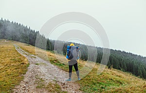 Male hiker with a backpack on his back standing on a trail in the mountains on a mountain meadow in rainy weather, view from the