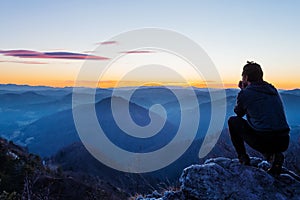 Male hiker crouching on top of the hill watching twilight landscape after sunset