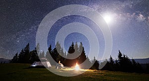 Male hiker enjoyng night camping near tourist tent at campfire under blue starry sky and Milky way
