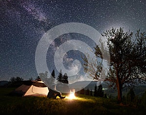 Male hiker enjoyng night camping near tourist tent at campfire under blue starry sky and Milky way