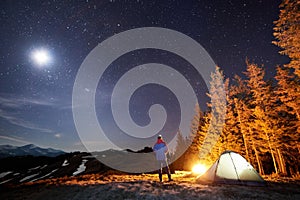 Male hiker have a rest in his camp near the forest at night under beautiful night sky full of stars and the moon