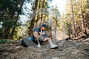 Male hiker in the mountains sitting by the trail with a bottle of water in his hand while relaxing and using a smartphone