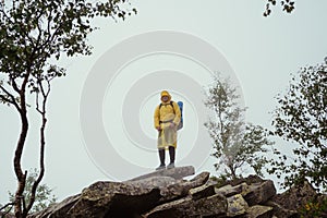 Male hiker in a raincoat with a backpack stands on a rock in the mountains during a hike and a foggy day