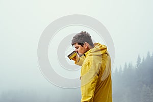 Male hiker in a yellow jacket drinks from a cup of hot tea on a background of misty view in the mountains