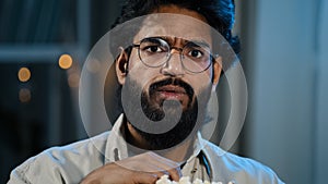 Male home portrait serious interested attentive Arabic Hispanic Indian bearded man guy in glasses eating popcorn