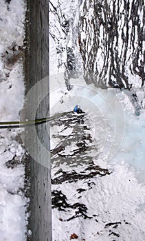 Male ice climber in a blue jacket rappelling a from a bridge railing onto a vertical frozen waterfall