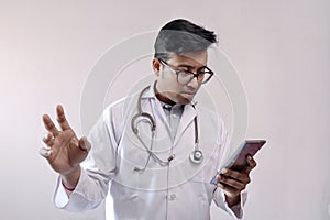 Male indian doctor in white coat and stethoscope in worried expression while looking at smart phone