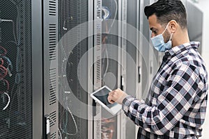 Male informatic engineer working inside server room database while wearing face mask during corona virus