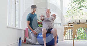 Male instructor kneeling by senior man exercising with dumbbells on fitness ball at gym