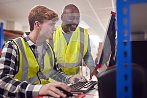 Male Intern With Supervisor Working In Busy Modern Warehouse On Computer Terminal