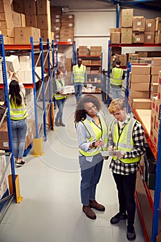 Male Intern With Team Leader Looking At Clipboard Inside Busy Warehouse Facility