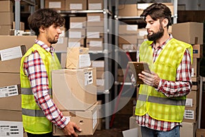 Male inventory manager shows digital tablet information to a arabic worker holding cardboard boxes, talking about work