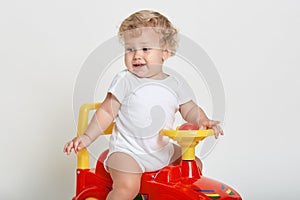 Male kid with wavy blond hair driving red and yellow toy car indoor, looking away, dresses white bodysuit, posing isolated over