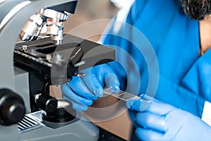 Male laboratory assistant examining biomaterial samples in a microscope. Cllose up hands in blue rubber gloves adjust microscope