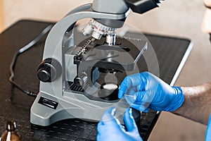 Male laboratory assistant examining biomaterial samples in a microscope. Cllose up hands in blue rubber gloves adjust microscope
