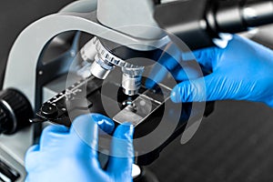 Male laboratory assistant examining biomaterial samples in a microscope. Close up hands in blue rubber gloves adjust microscope