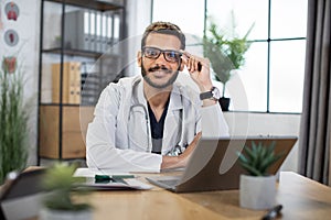 Male Malay doctor in eyeglasses, sitting at the table with laptop pc in modern office