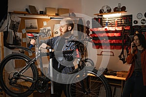 Male mechanic repairing a bicycle in a workshop or garage, his woman waiting in the background. Bike Maintenance