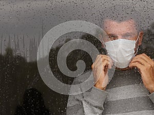 Male in medical mask looking through window in a sad rainy day.