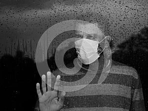 Male in medical mask looking through window in a sad rainy day.