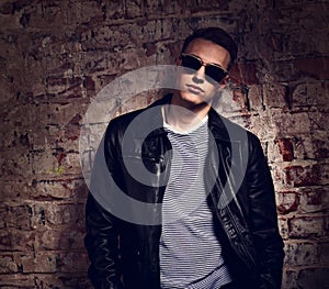 Male model posing in leather jacket and trendy eyeglasses on brick wall street background. fashion toned contrast portrait with e