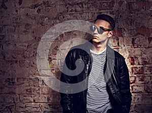 Male model posing in leather jacket and trendy eyeglasses on brick wall street background. Fashion toned contrast portrait with e