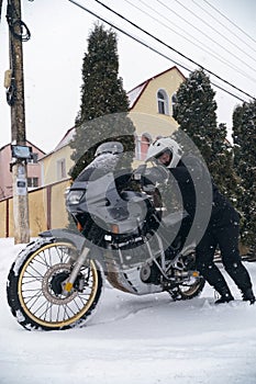 A male motorcyclist in a special outfit pushes his motorcycle on a snowy slippery road. Winter fun, stupid idea