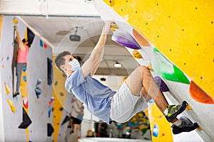Male mountaineer in protective mask climbing artificial rock wall without his belay