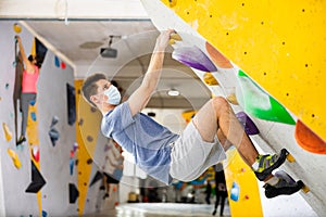 Male mountaineer in protective mask climbing artificial rock wall without his belay