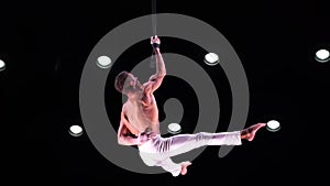 Male, muscular circus artist on Aerial straps on black stage background performing gymnastics trick. Concept of