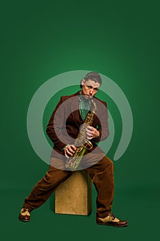 Male musician in brown classical suit playing saxophone against dark green studio background. Jazz music concert