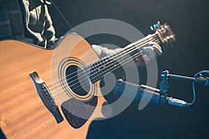 Male musician playing acoustic guitar behind microphone in recording studio.