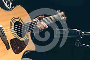 Male musician playing acoustic guitar behind microphone in recording studio.