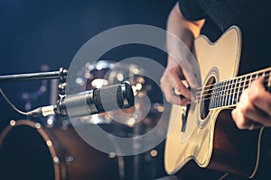 Male musician playing acoustic guitar behind microphone in recording studio.