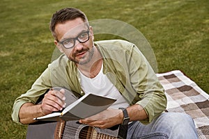 Male musician working in park. Young focused man wearing eyeglasses holding acoustic guitar and composing a song while