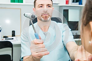 Male neurologist doctor inspecting young female patient`s nervous system using a hammer. Neurological physical examination. Helth