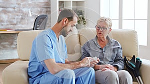 Male nurse sitting on couch with senior woman giving her medical treatment in nursing home