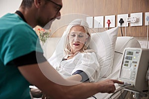 Male Nurse Taking Mature Female Patients Blood Pressure In Hospital Bed With Automated Machine