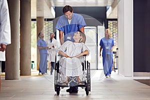 Male Nurse Wearing Scrubs Pushing Female Patient In Wheelchair Through Hospital Building