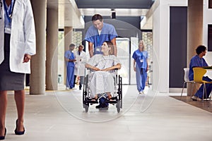 Male Nurse Wearing Scrubs Pushing Female Patient In Wheelchair Through Hospital Building