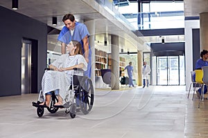 Male Nurse Wearing Scrubs Pushing Female Patient In Wheelchair Through Hospital Building