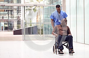 Male Nurse Wearing Scrubs Wheeling Patient In Wheelchair Through Lobby Of Modern Hospital Building