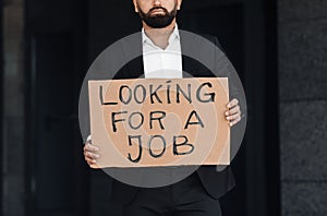 Male office worker in suit holding cardboard sign written looking for a job text, standing outside business center