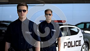 Male officers in sunglasses standing against squad car, ready for patrolling
