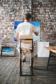 Male painter sits on chair in front canvas and drawing picture in studio. Art class and workshop. Artist painting process. Hobby