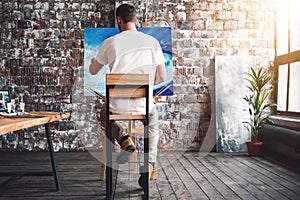 Male painter sits on chair in front canvas and drawing picture in studio. Art class and workshop. Artist painting process